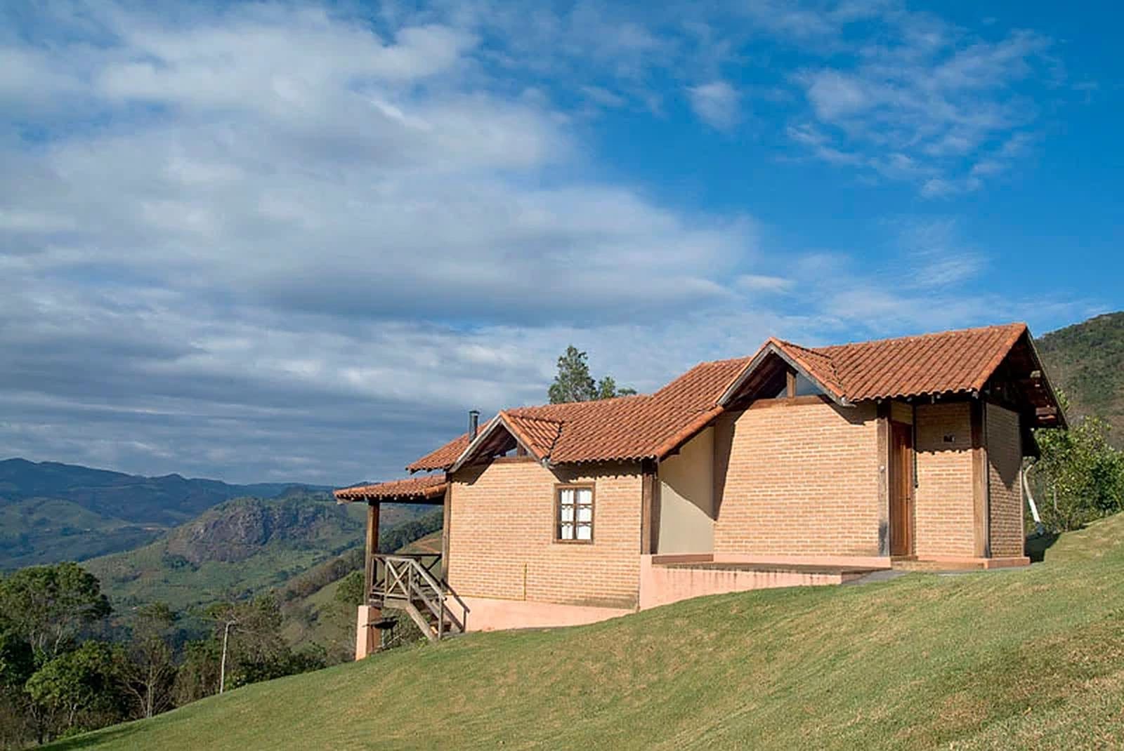 Chalé de tijolos sobre o gramado, com montanhas e céu azul ao fundo